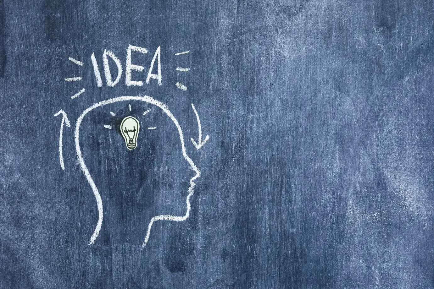 Лампочка-символ идеи внутри нарисованного контура лица, с текстом «idea» на меловой доске.